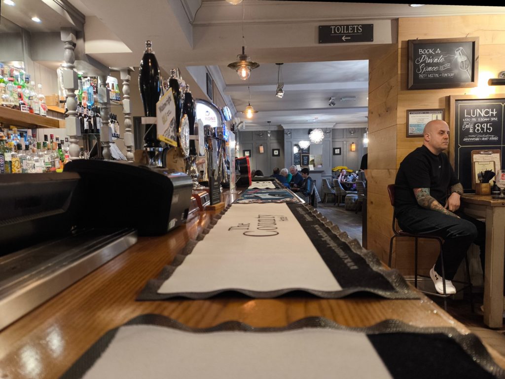 Looking along the bar at the County Hotel in Hexham with the bar to the left and drinkers to the right with the dining area in the background
