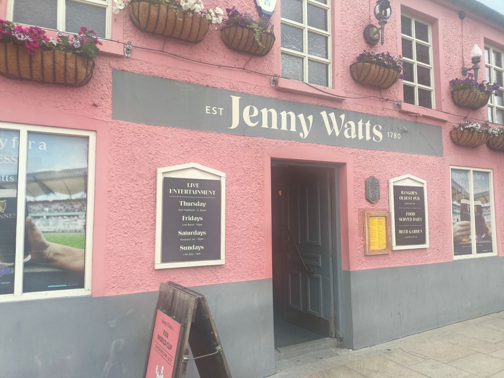Exterior of The Jenny Watts pub in Bangor showing the hanging baskets, pub signage and the pink painted frontage.
